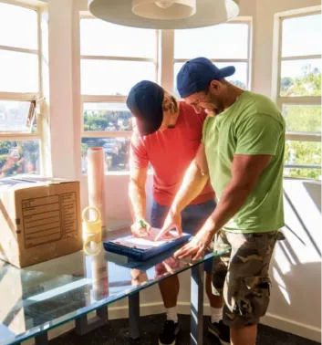 Two people in casual clothing, one in a red shirt and the other in a green shirt, are writing on a clipboard in a well-lit room with large windows. A cardboard box and packing tape sit on a glass table, as they prepare for their long distance moving service.