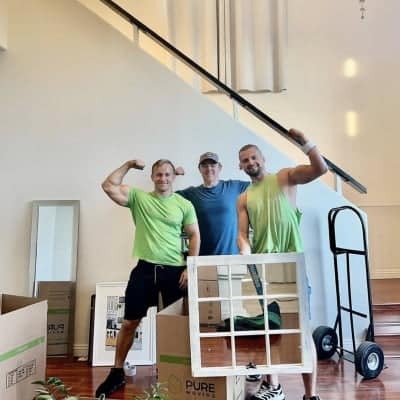 Three men, one in a blue shirt and two in green shirts, pose while flexing their arms in a partially furnished room with moving boxes, a hand truck nearby, and competitive moving rates.